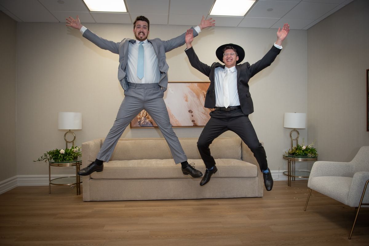 Two men in suits leap joyfully off a beige sofa in a modern room with bright lighting and decorative elements.