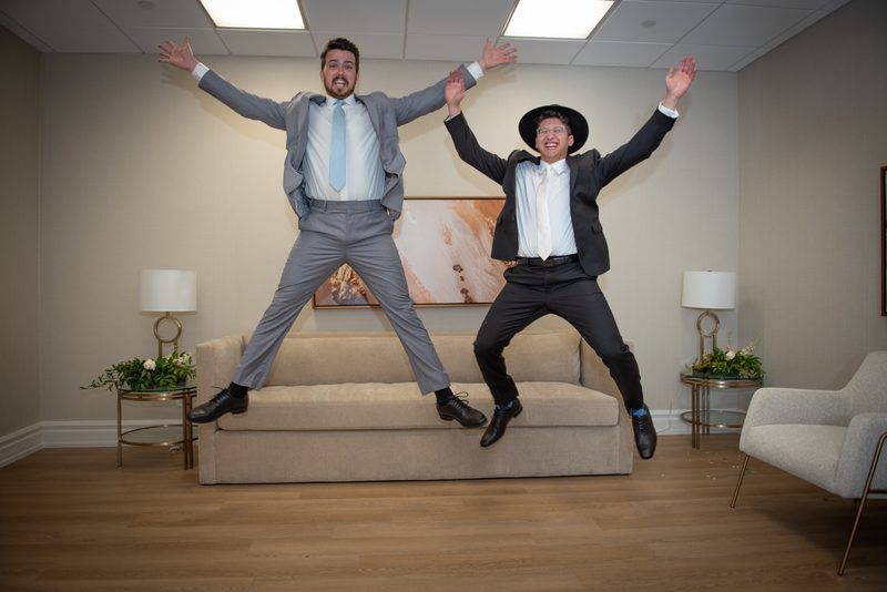 Two men in suits leap joyfully off a beige sofa in a modern room with bright lighting and decorative elements.