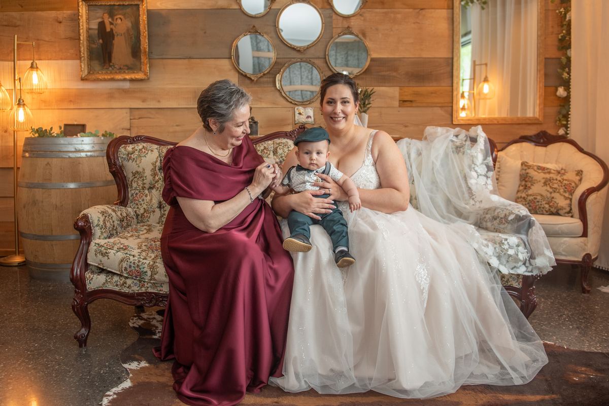 A bride holds a baby while an older woman in burgundy interacts with the child, creating a warm family moment.