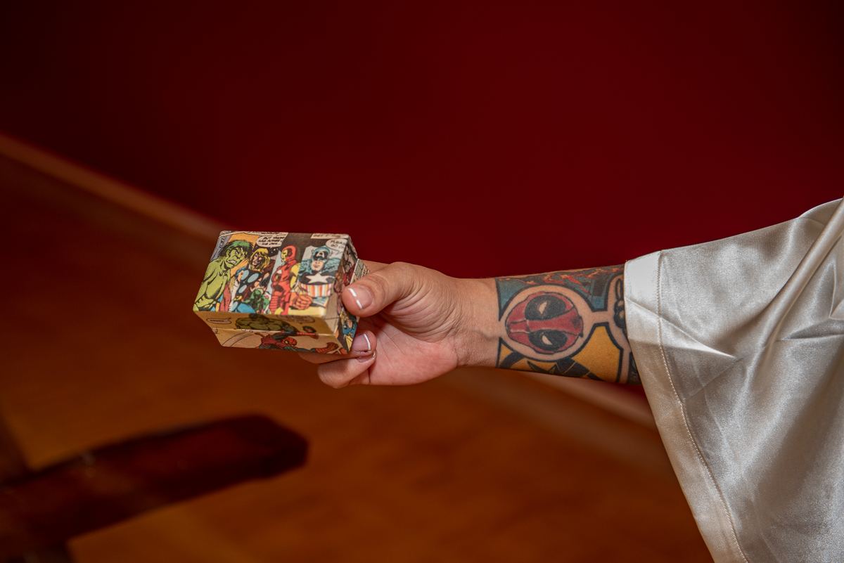 A person with a Deadpool tattoo holds up a comic-book-patterned box in front of a deep red backdrop.