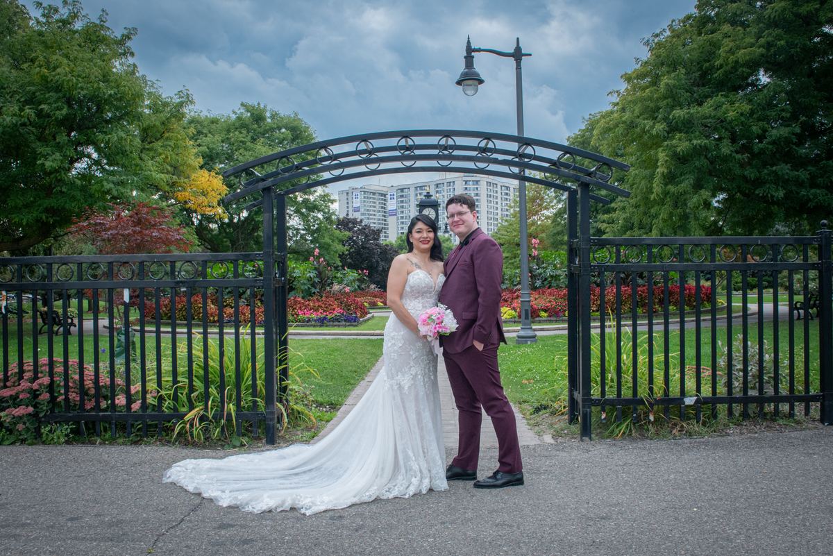 A newlywed couple in formal attire stands smiling beneath an ornate black metal archway surrounded by greenery and flowers.