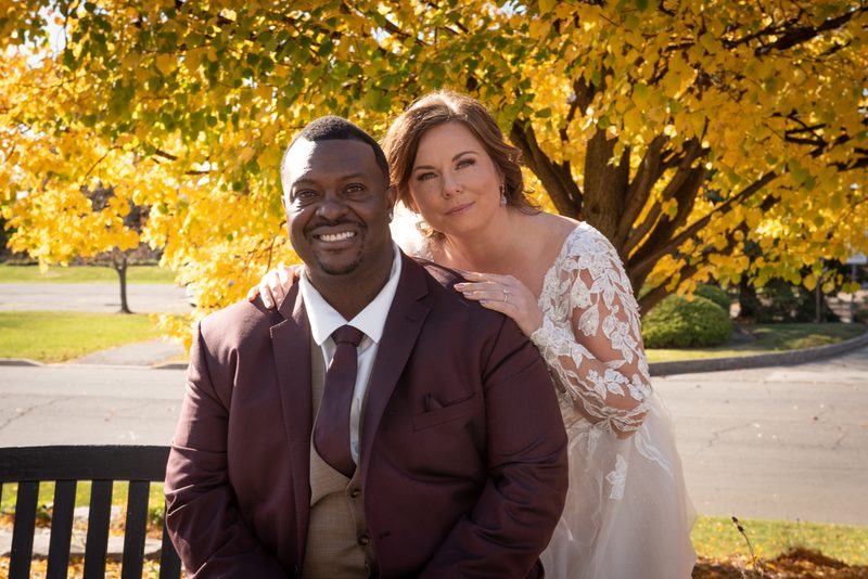 A newlywed couple in formal attire stands under a canopy of golden autumn leaves, sunlight filtering through the foliage.