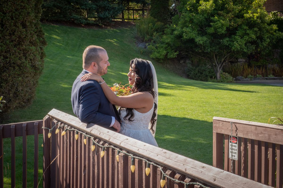 A bride and groom embrace tenderly on a wooden deck, overlooking a sunlit lawn with string lights hanging along the railing.