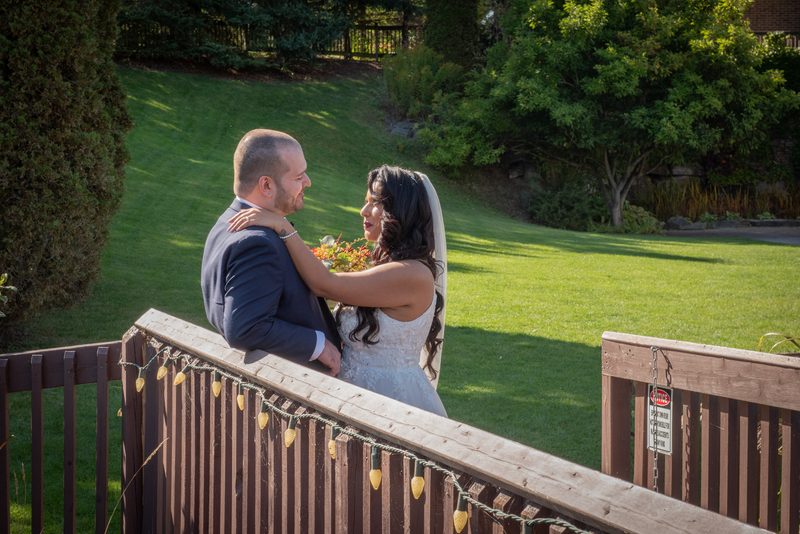 A bride and groom embrace tenderly on a wooden deck, overlooking a sunlit lawn with string lights hanging along the railing.