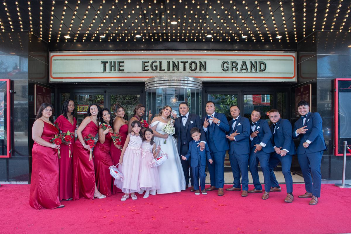 A wedding party poses joyfully on a red carpet in front of a lit cinema marquee, smiling and striking playful poses.