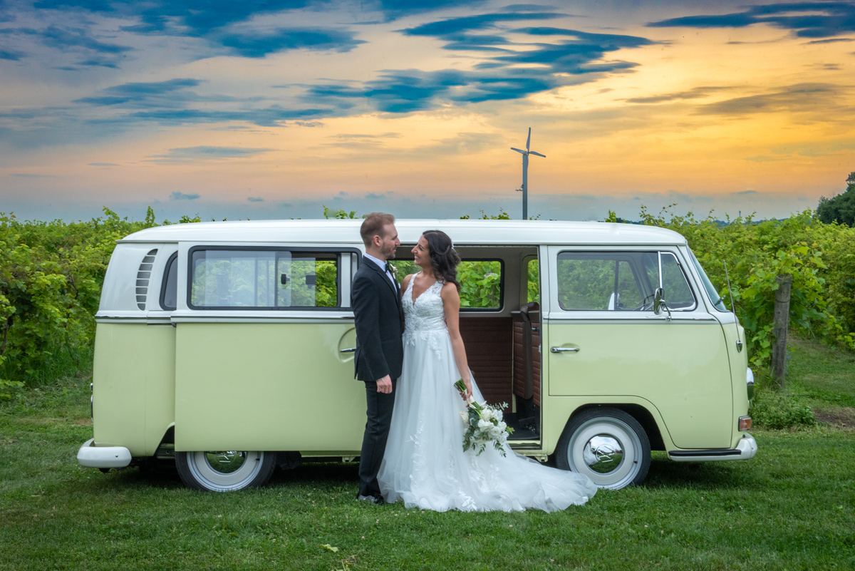 A newlywed couple in wedding attire embrace beside a vintage Volkswagen van at sunset in a vineyard.