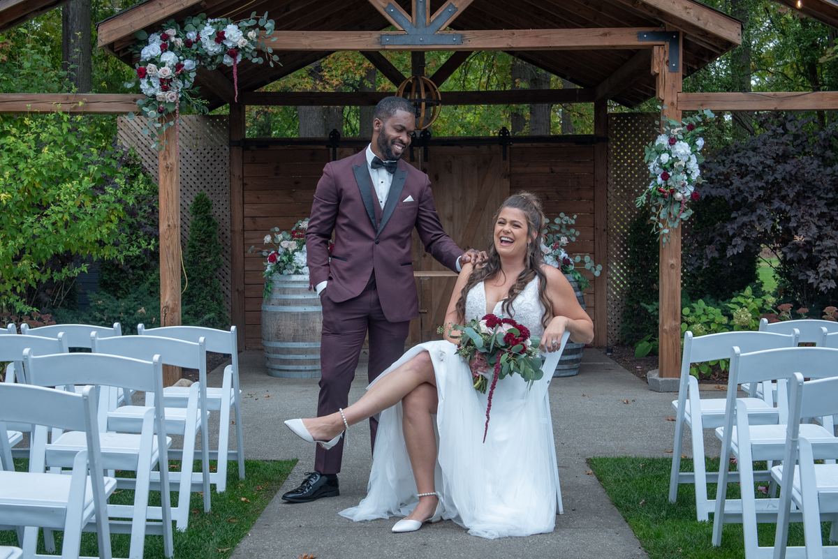 Bride and groom laugh together under a floral-adorned wooden arbor during an outdoor wedding ceremony.