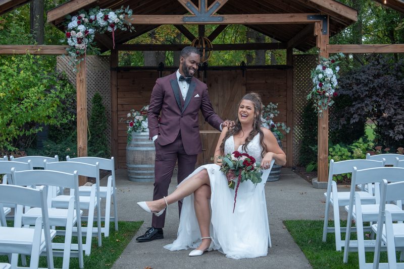 Bride and groom laugh together under a floral-adorned wooden arbor during an outdoor wedding ceremony.