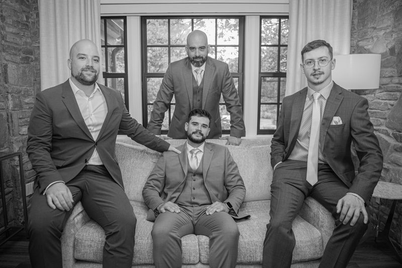 Four men in suits and ties stand around a sofa in a stone-walled room bathed in soft natural light from large windows.