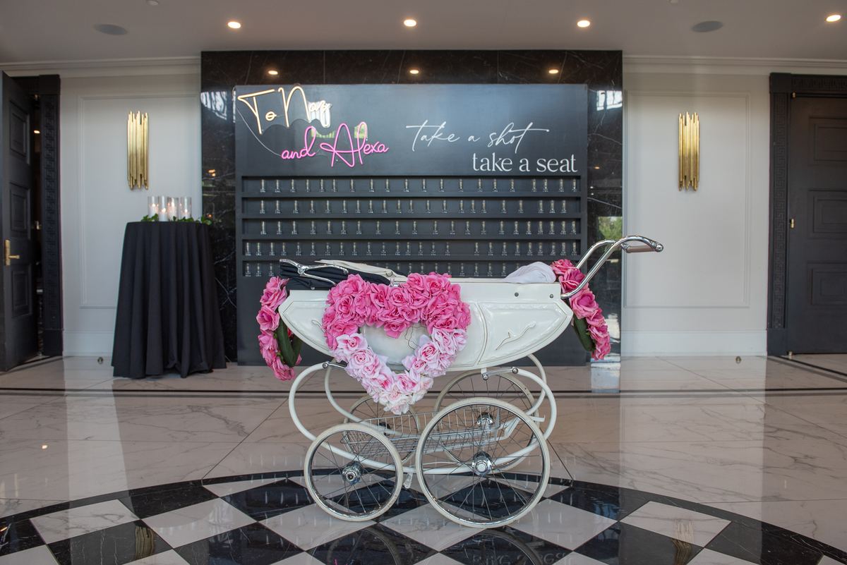 A vintage white baby carriage decorated with a pink rose heart in front of a neon sign on a black wall at an event space.