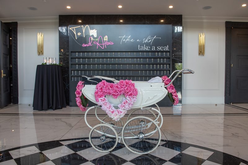 A vintage white baby carriage decorated with a pink rose heart in front of a neon sign on a black wall at an event space.