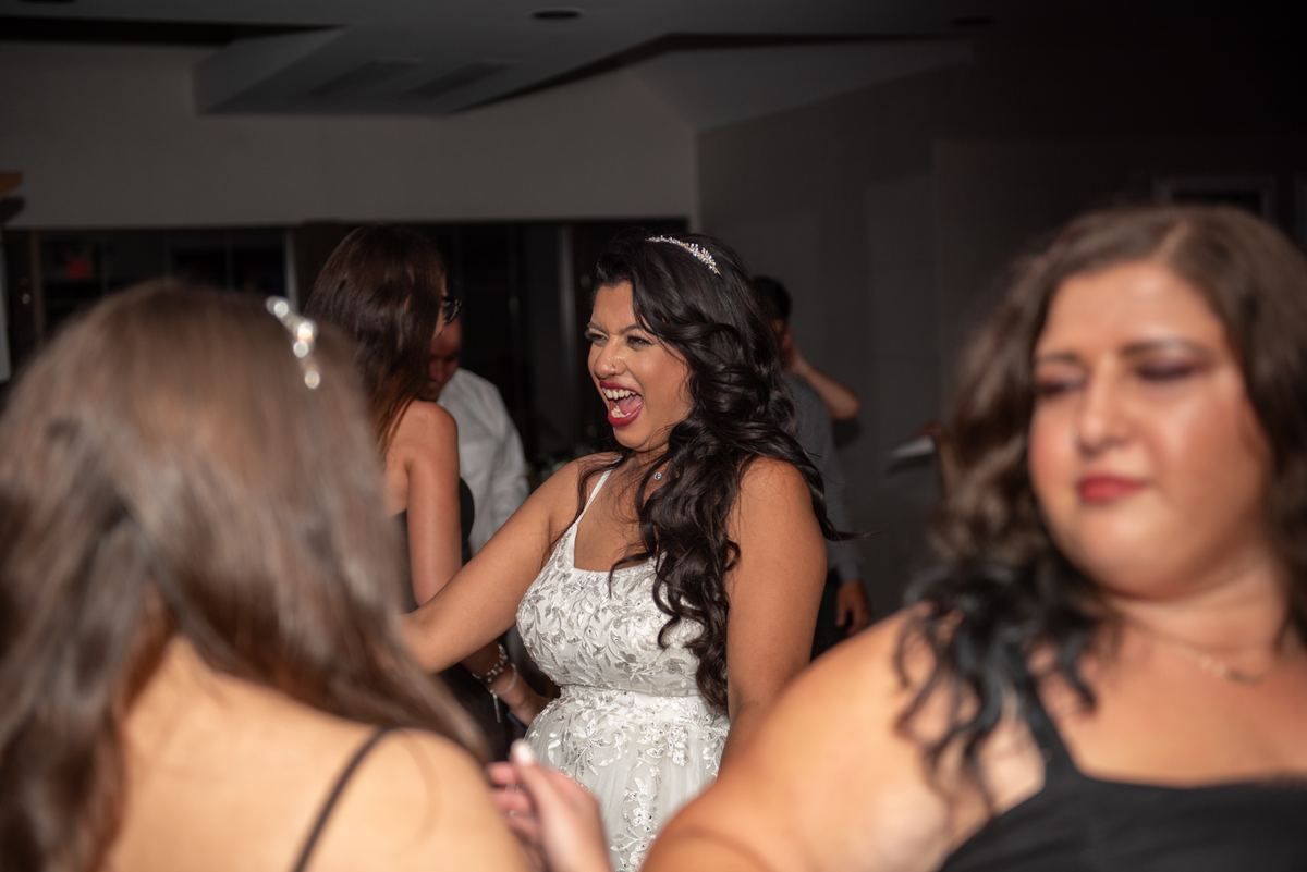 A laughing bride in a white lace gown stands out at a lively reception, surrounded by blurred guests and warm lighting.
