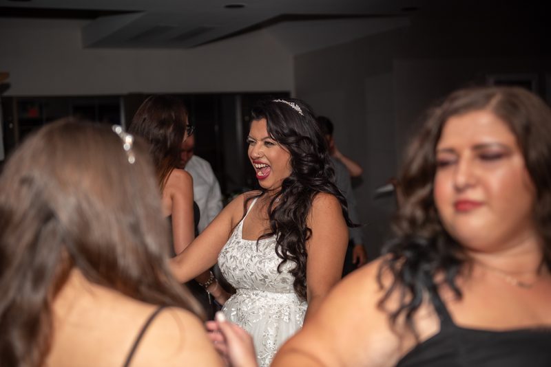 A laughing bride in a white lace gown stands out at a lively reception, surrounded by blurred guests and warm lighting.