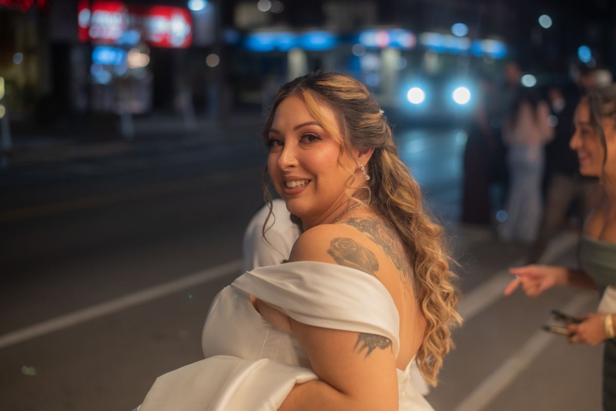 A bride in a white off-the-shoulder gown walks through a nighttime urban setting, smiling and looking back over her shoulder.