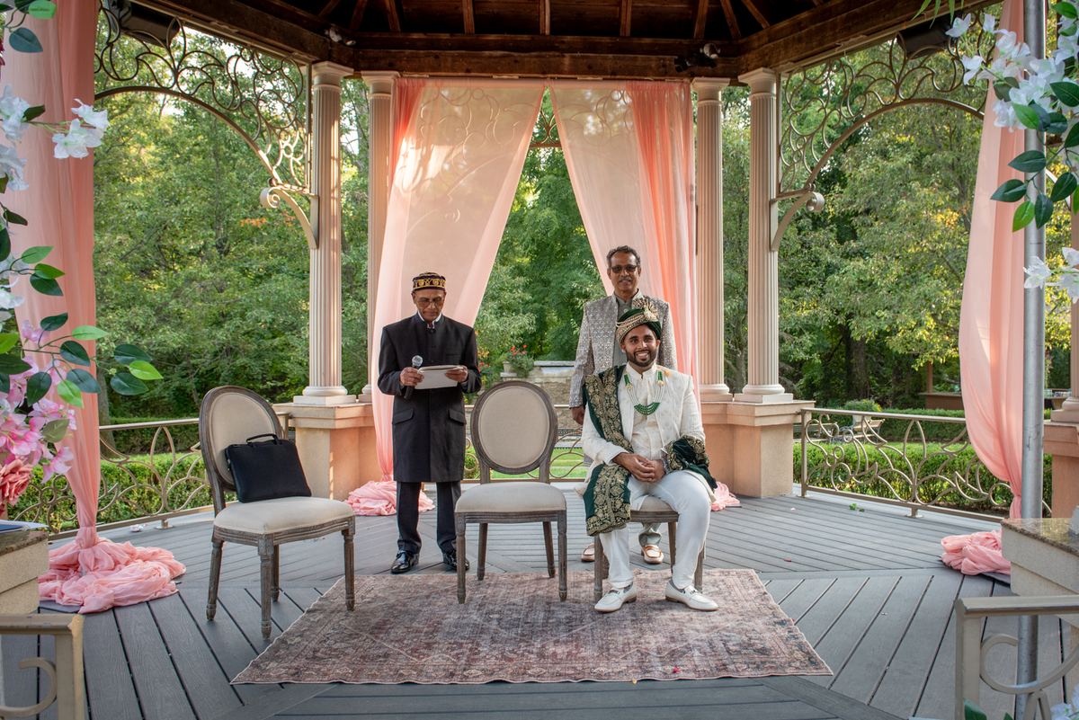 A groom in white and green sits smiling during a wedding ceremony under an ornate gazebo with pink drapes.