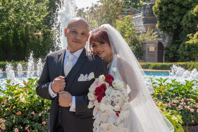 A newlywed couple in formal attire stands before a fountain in a sunlit garden filled with red and white roses.