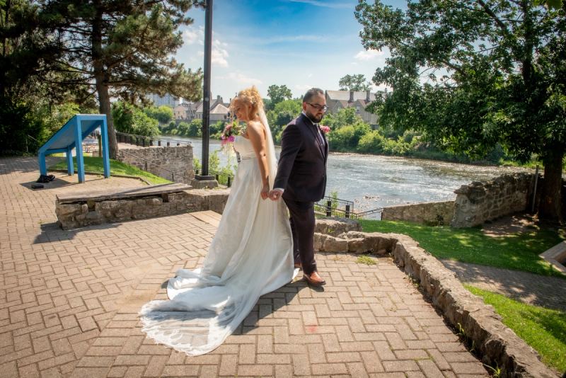 A smiling bride and groom stroll along a stone riverside path, sunlight highlighting the lush greenery.