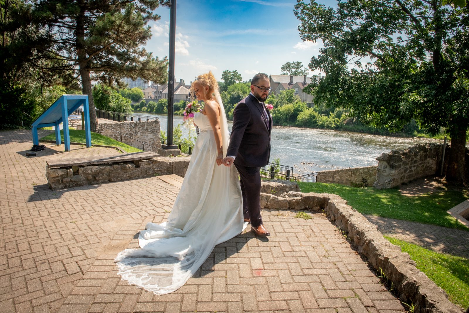 A smiling bride and groom stroll along a stone riverside path, sunlight highlighting the lush greenery.