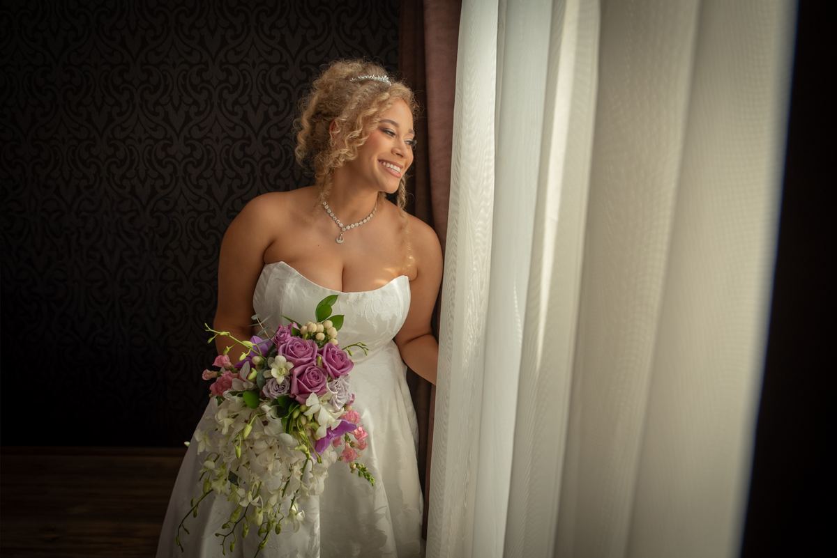 A bride in a white gown with a purple and white flower bouquet stands by a window, smiling at the view.