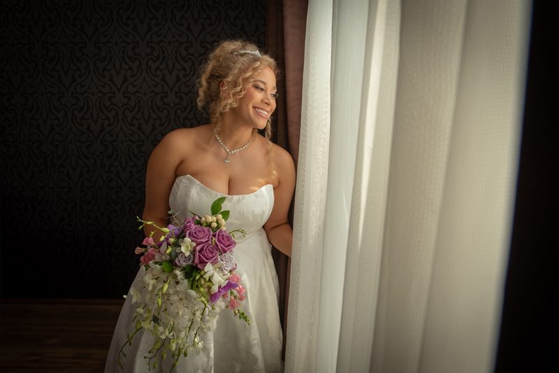 A bride in a white gown with a purple and white flower bouquet stands by a window, smiling at the view.