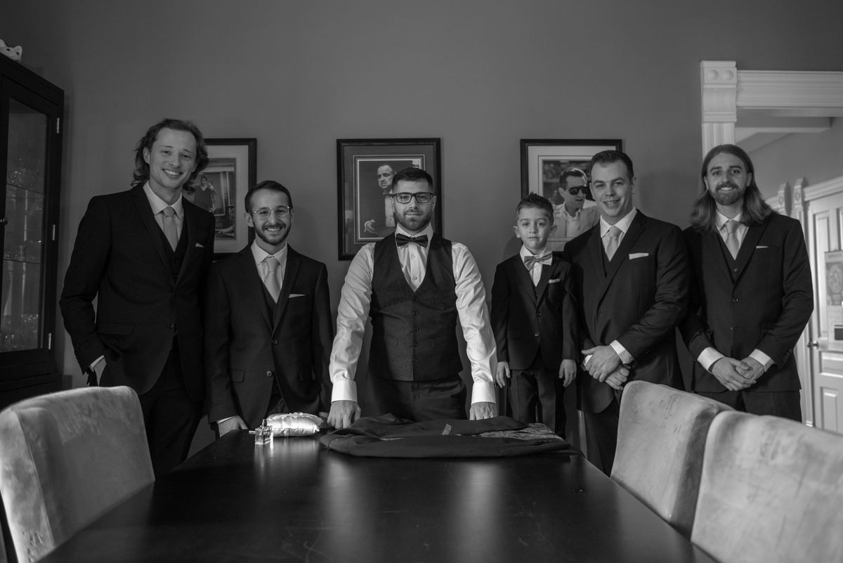 Six formally dressed men and a young boy stand around a table in a warmly lit room, suggesting a wedding gathering.