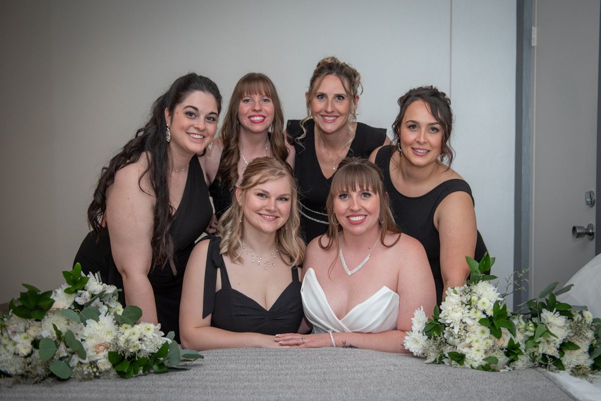 Bride in white gown surrounded by smiling bridesmaids in black dresses, framed by floral arrangements.