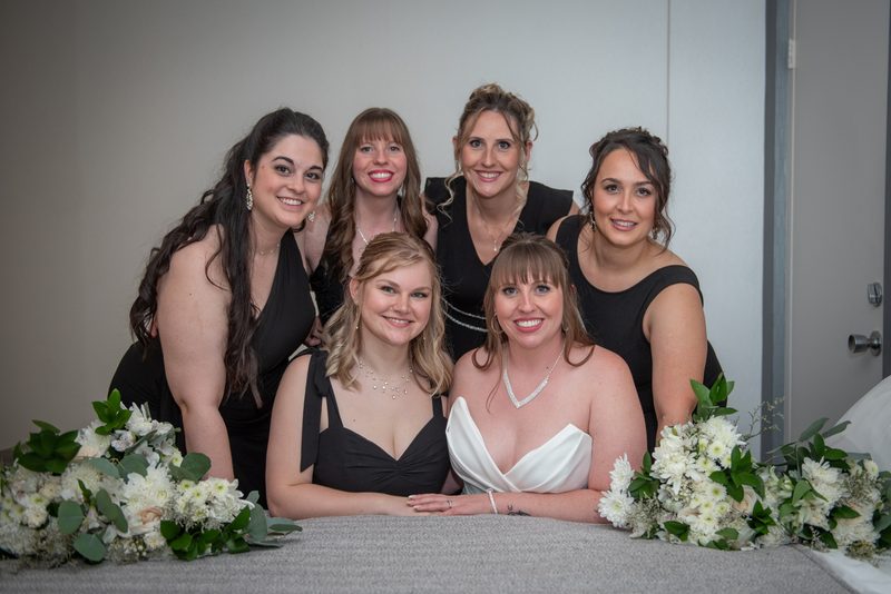 Bride in white gown surrounded by smiling bridesmaids in black dresses, framed by floral arrangements.