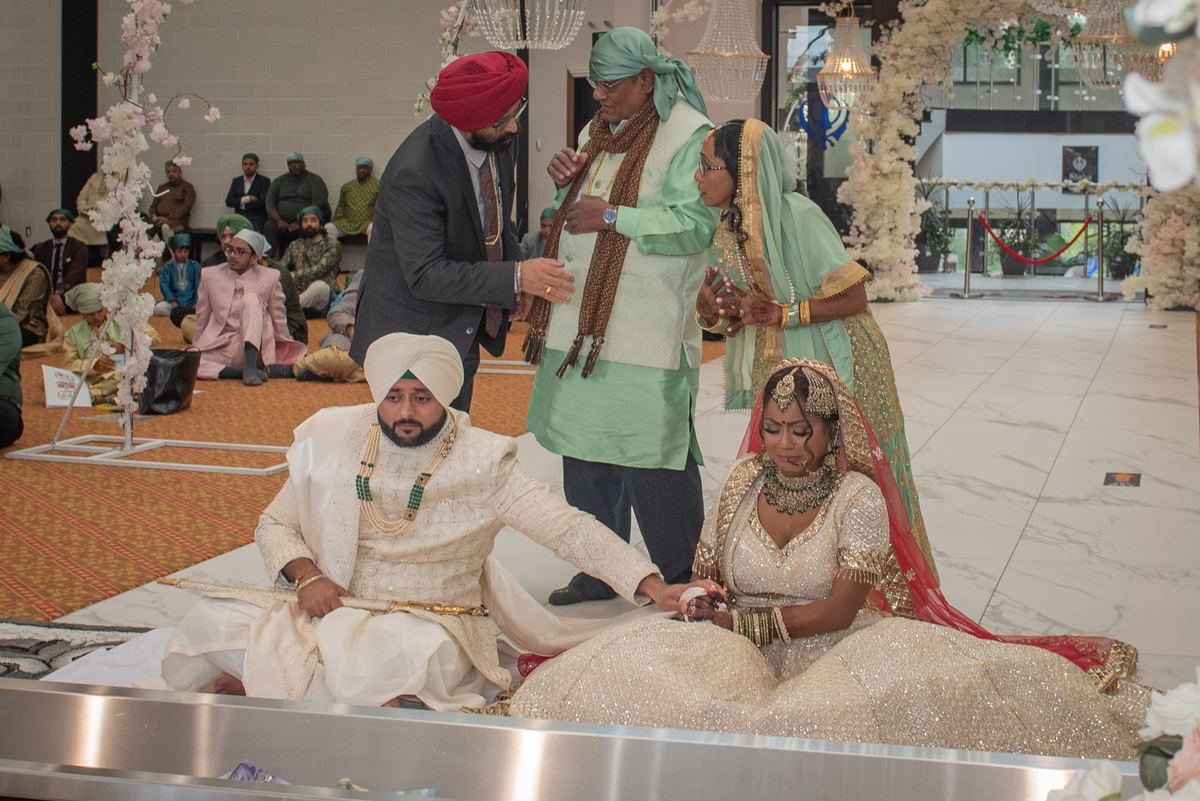 A Sikh couple exchange vows in a decorated hall surrounded by family and guests in traditional attire.