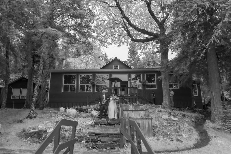 A bride and groom stand on stone steps in front of a cabin surrounded by trees as guests watch from the porch.