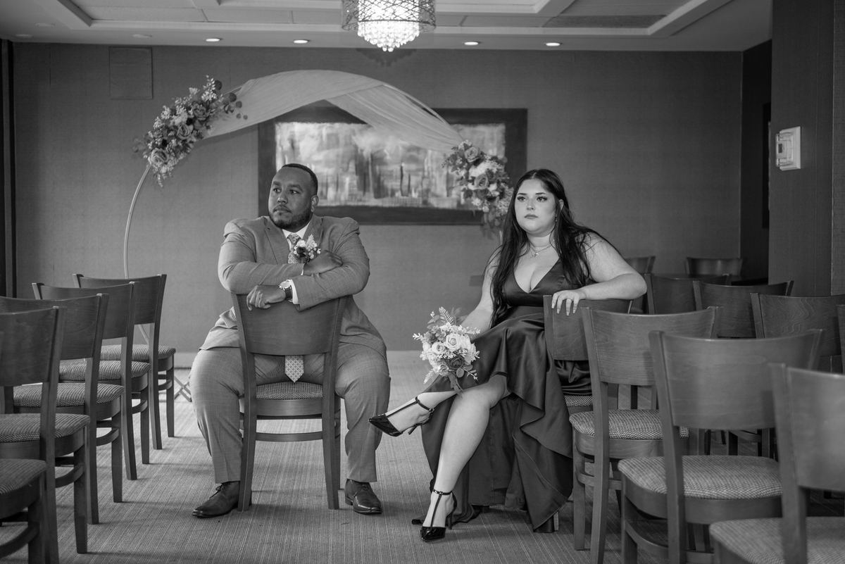 A formally dressed couple awaits a wedding ceremony in softly lit rows of chairs, framed by an arched floral decoration.