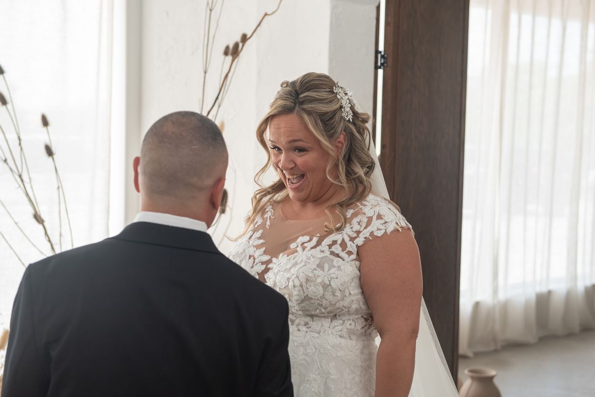 Bride in lace gown with floral hairpiece stands beside groom in dark suit, looking joyful during wedding ceremony.