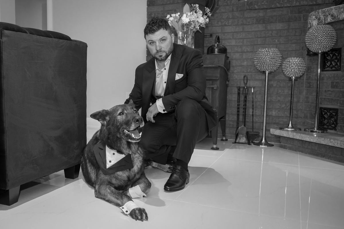 A man and his dog, both in formal tuxedos, pose playfully in a modern interior with glossy floors and soft lighting.
