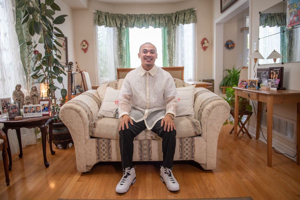 A man in a white barong tagalog and sneakers sits on a patterned sofa in a cozy living room with natural light.