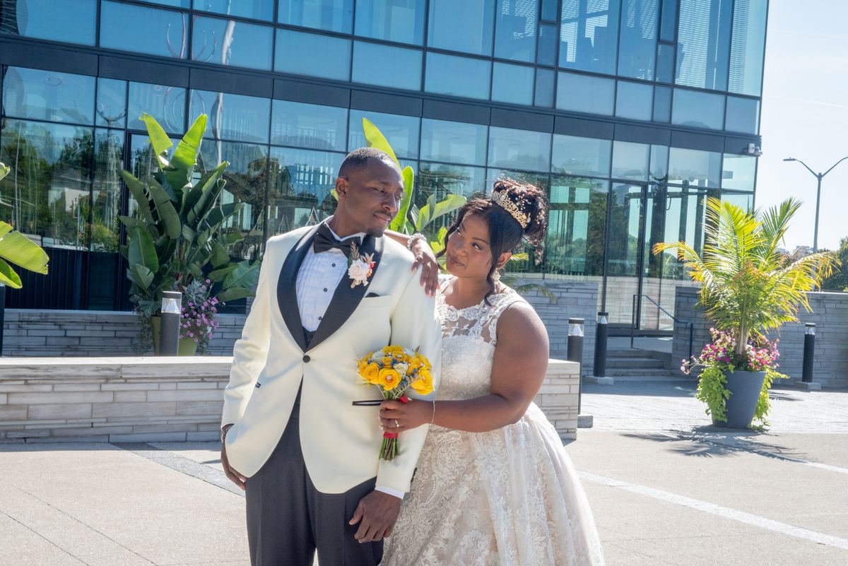 A groom in a cream tuxedo and his bride with yellow flowers stand outdoors near a modern glass building.