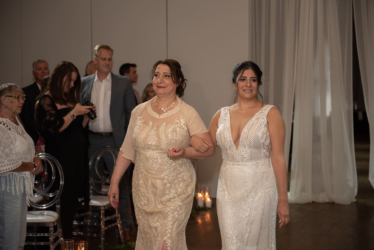 A bride in a white lace gown walks down the aisle with a family member, surrounded by warm candlelight and drapery.