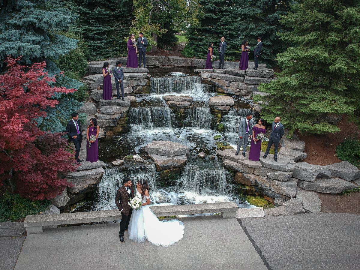 A bride and groom embrace in a serene stone waterfall garden surrounded by their purple-clad bridal party.