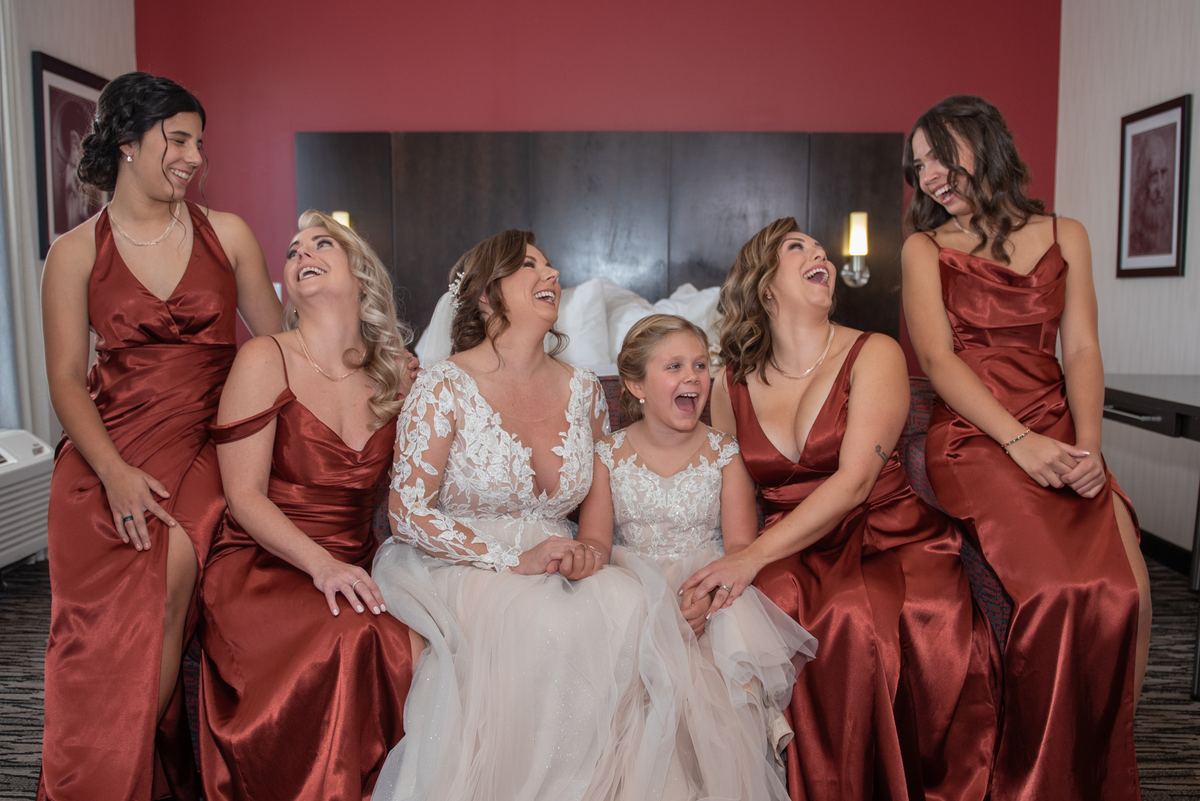 A bride, bridesmaids in rust satin, and a flower girl in white lace laugh together by bedside lamps in a hotel room bed.
