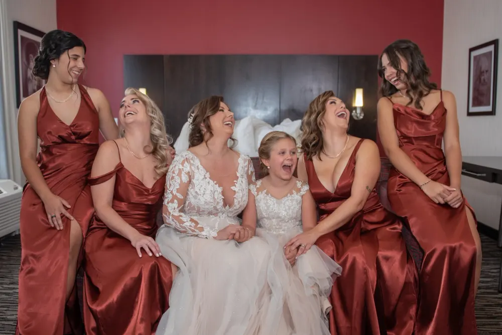 A bride, bridesmaids in rust satin, and a flower girl in white lace laugh together by bedside lamps in a hotel room bed.