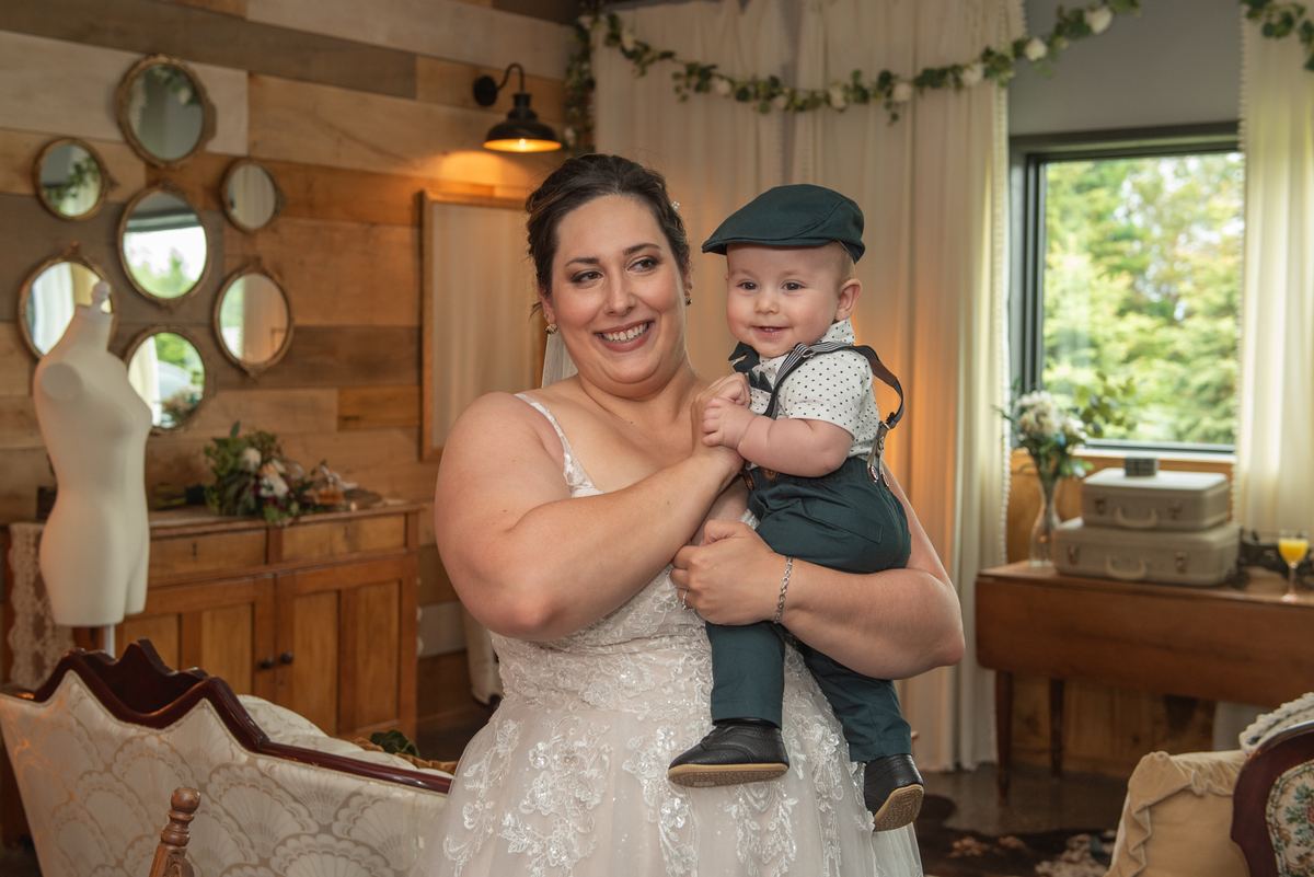 A bride and her baby boy smile at the camera in a rustic room with wooden paneling and floral garlands.