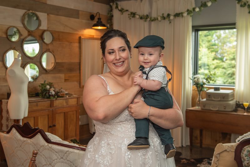 A bride and her baby boy smile at the camera in a rustic room with wooden paneling and floral garlands.