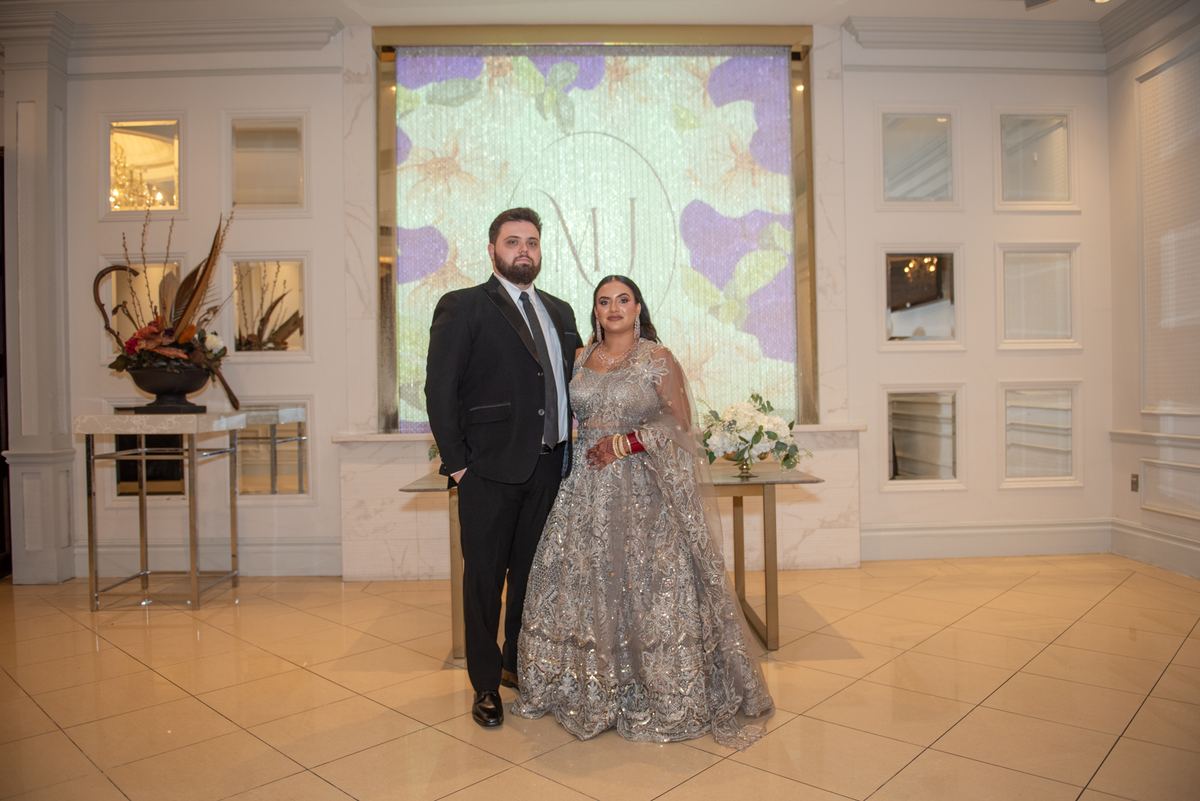 A bride in a silver lehenga and groom in a black suit pose with an illuminated monogram backdrop, surrounded by flowers.
