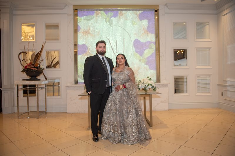 A bride in a silver lehenga and groom in a black suit pose with an illuminated monogram backdrop, surrounded by flowers.