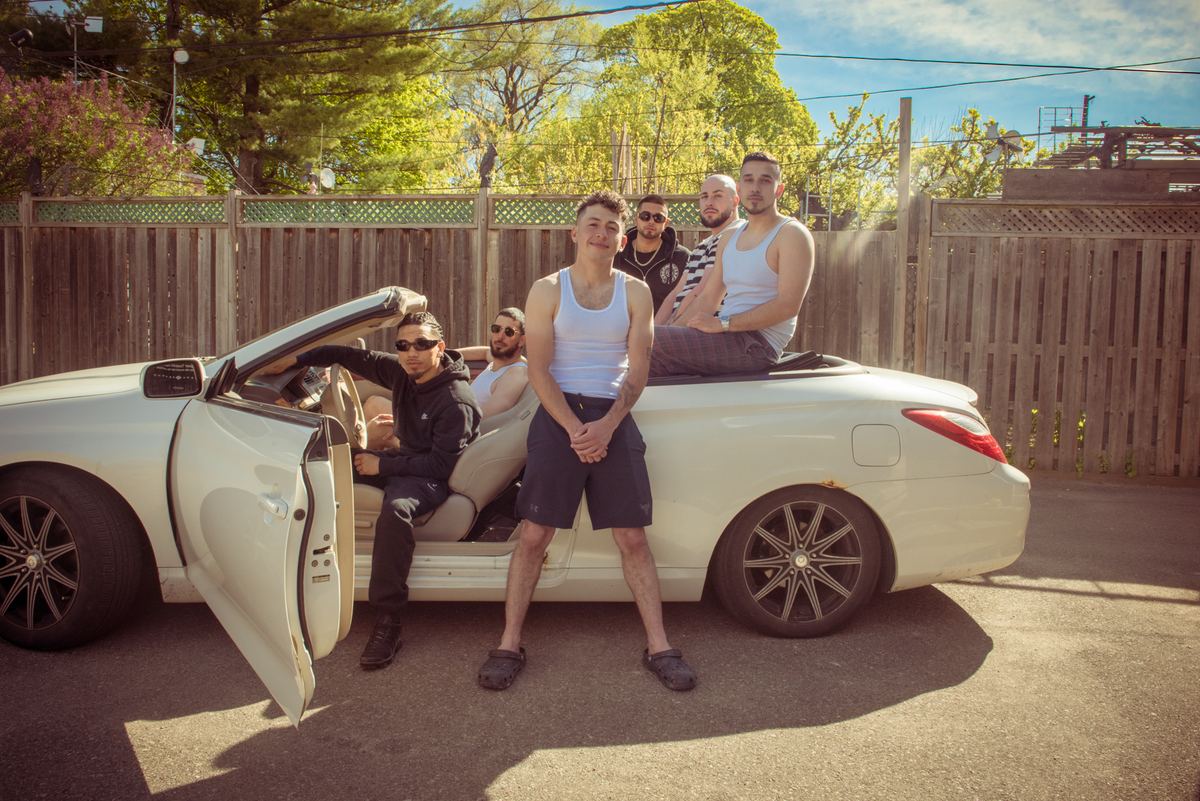 Six young men pose casually around a cream-colored convertible in afternoon sunlight, exuding urban camaraderie.