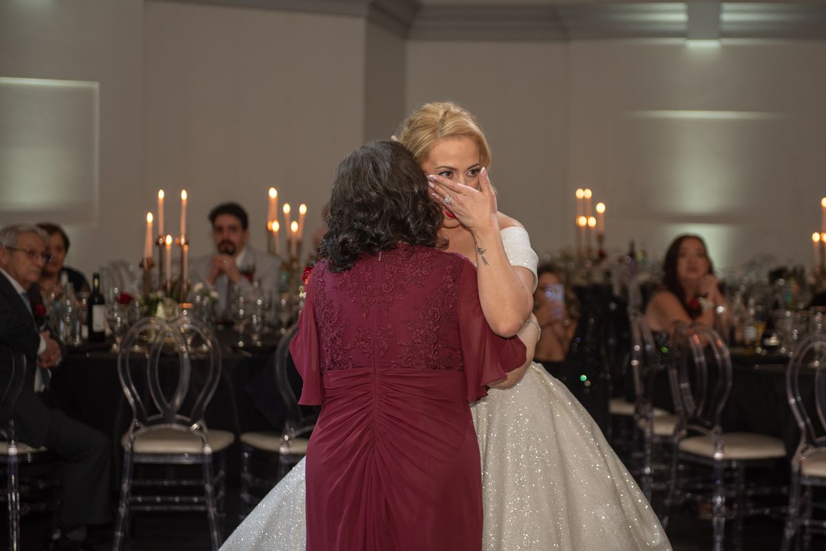 A bride in a sparkling gown embraces her emotional mother, who covers her face with her hand at the reception.