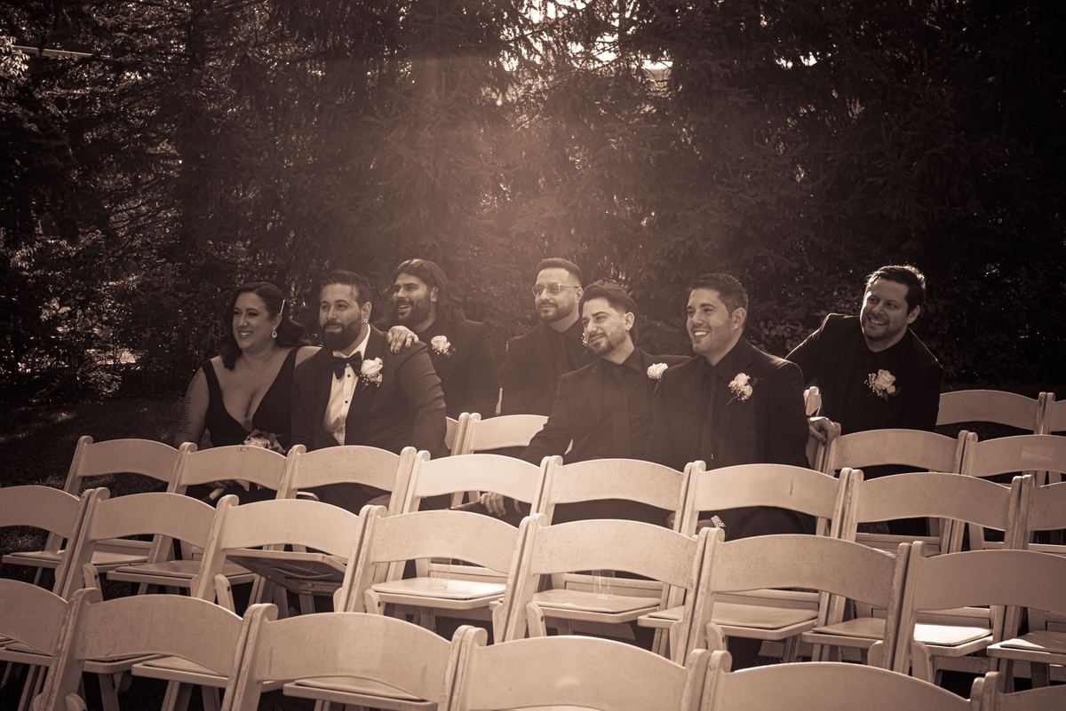 A joyful wedding party in wooden chairs outdoors, bathed in warm golden light filtering through trees.