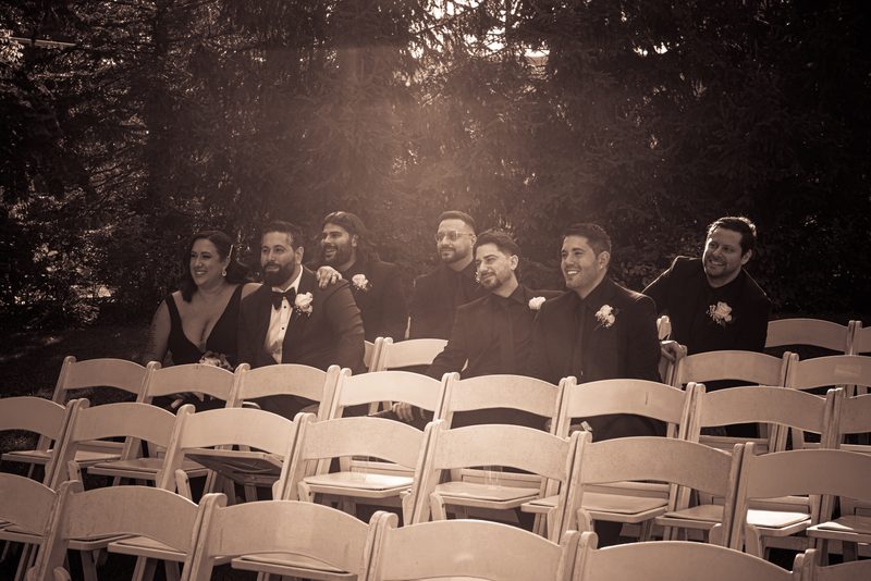A joyful wedding party in wooden chairs outdoors, bathed in warm golden light filtering through trees.