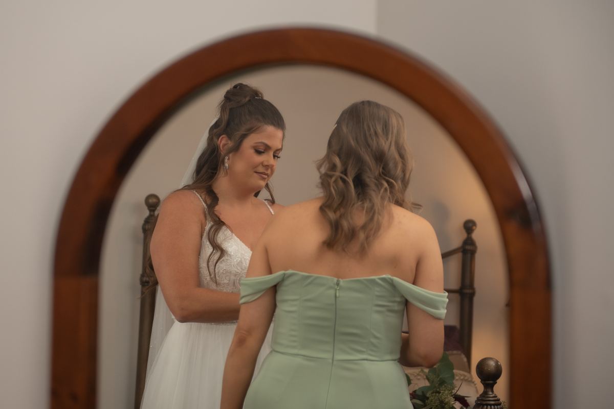 A bride in a white gown helps adjust her bridesmaid's sage-green off-the-shoulder dress near an arched mirror.