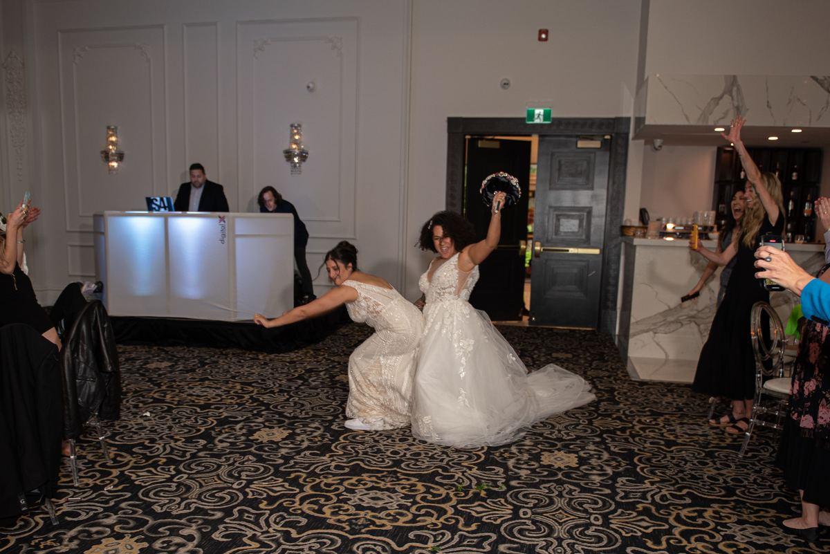 Two brides playfully tug on a ribbon, one raising her bouquet as guests cheer around them in an event space.