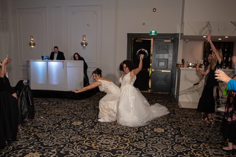 Two brides playfully tug on a ribbon, one raising her bouquet as guests cheer around them in an event space.