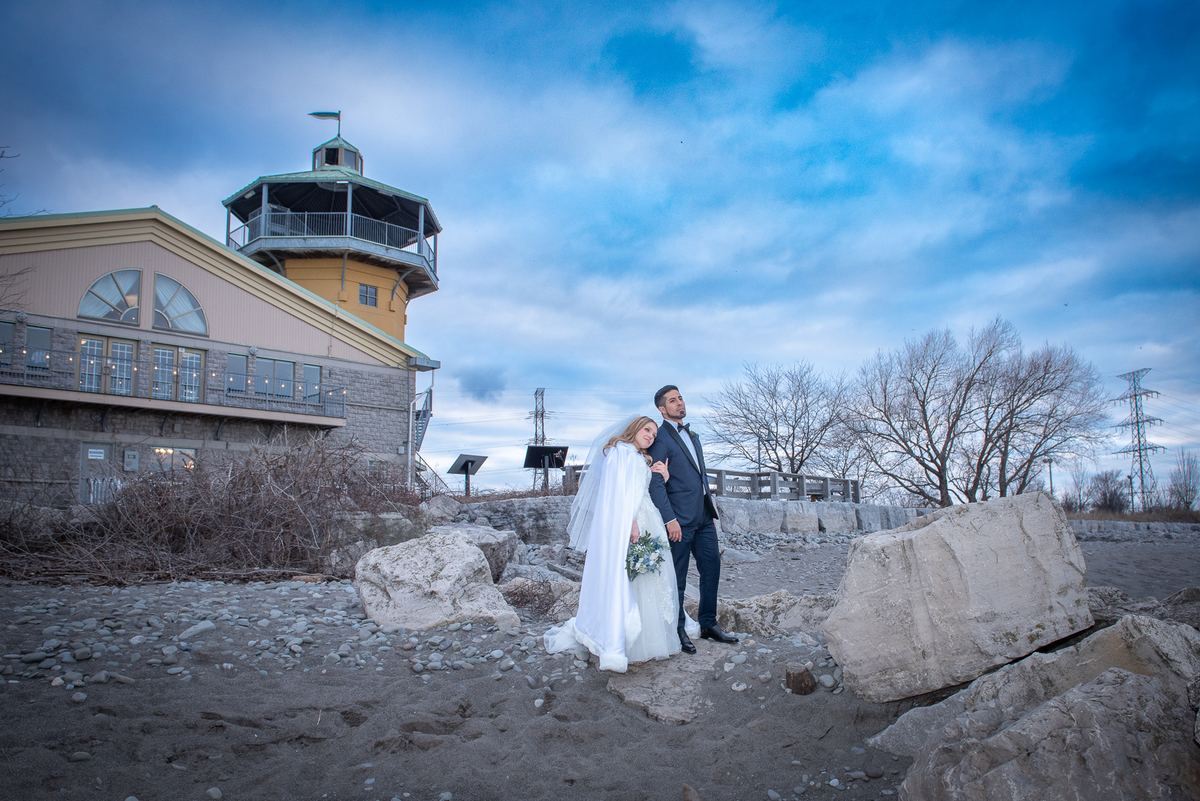 A bride and groom stand on a rocky shoreline with a towered building in the background under a dramatic blue and cloudy sky.